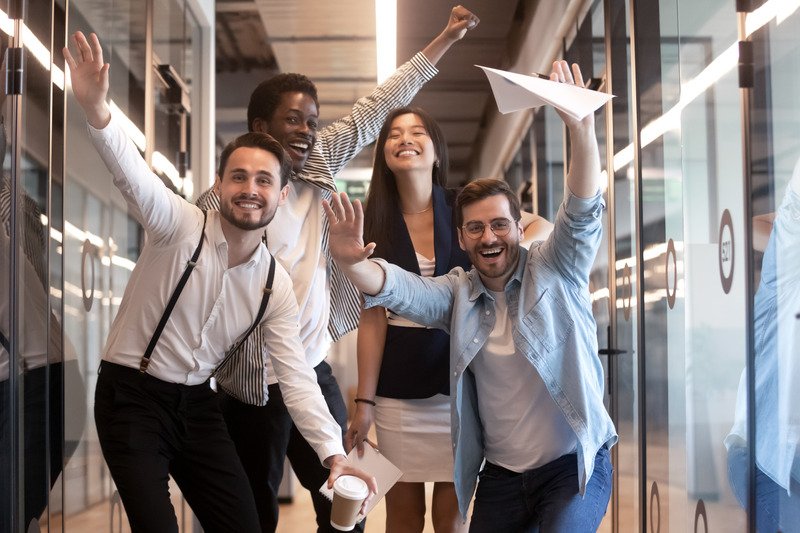 Five colleagues cheering with their arms raised as they walk down a glass-walled office corridor