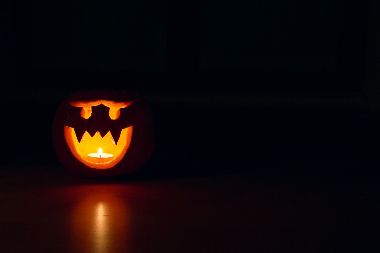 A carved jack-o’-lantern lit by a single tea light, sitting alone on the floor of a dark room, its glow reflected on the boards in front of it