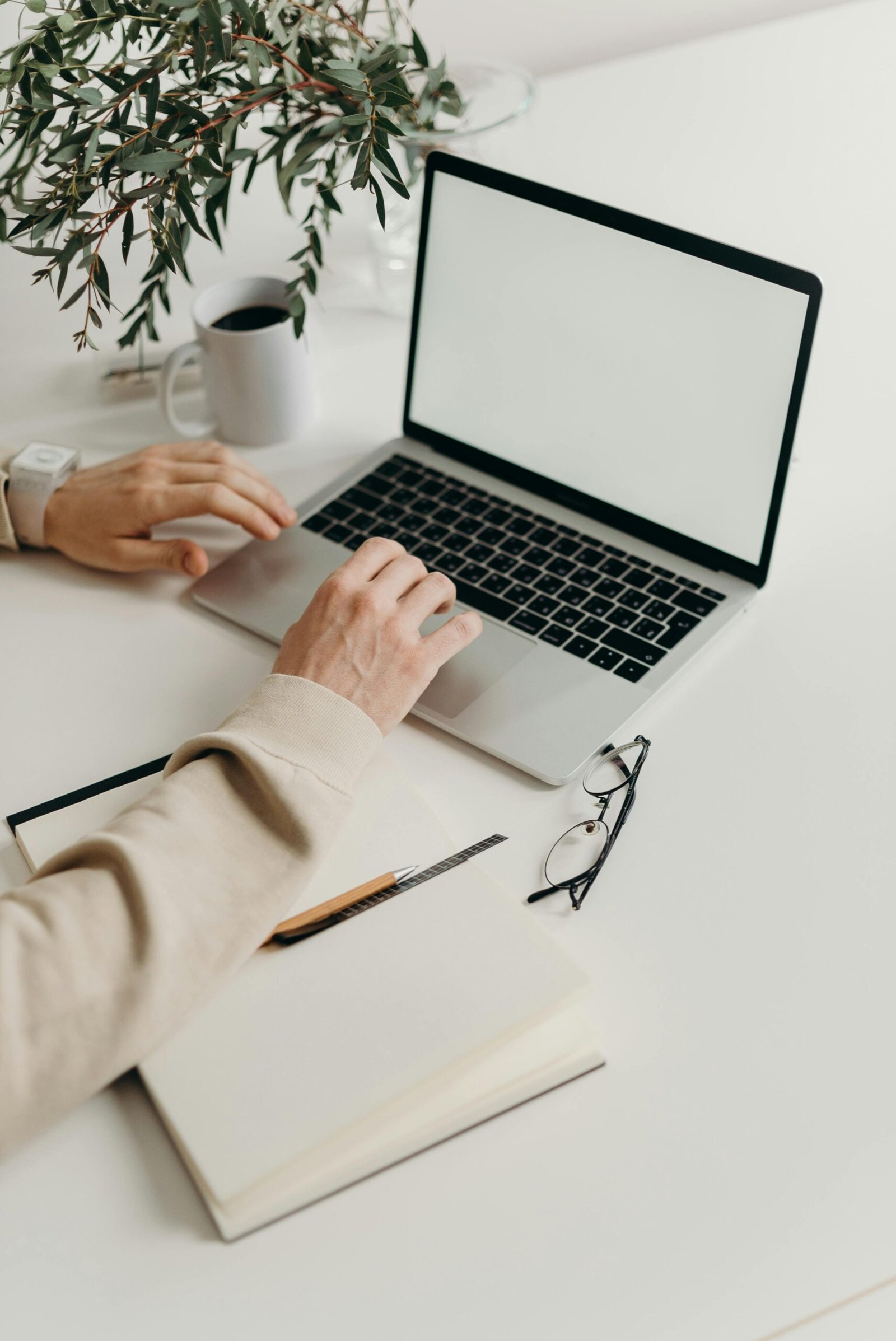 Someone working at a laptop on a white desk beside a notebook, pen, glasses, and a cup of coffee