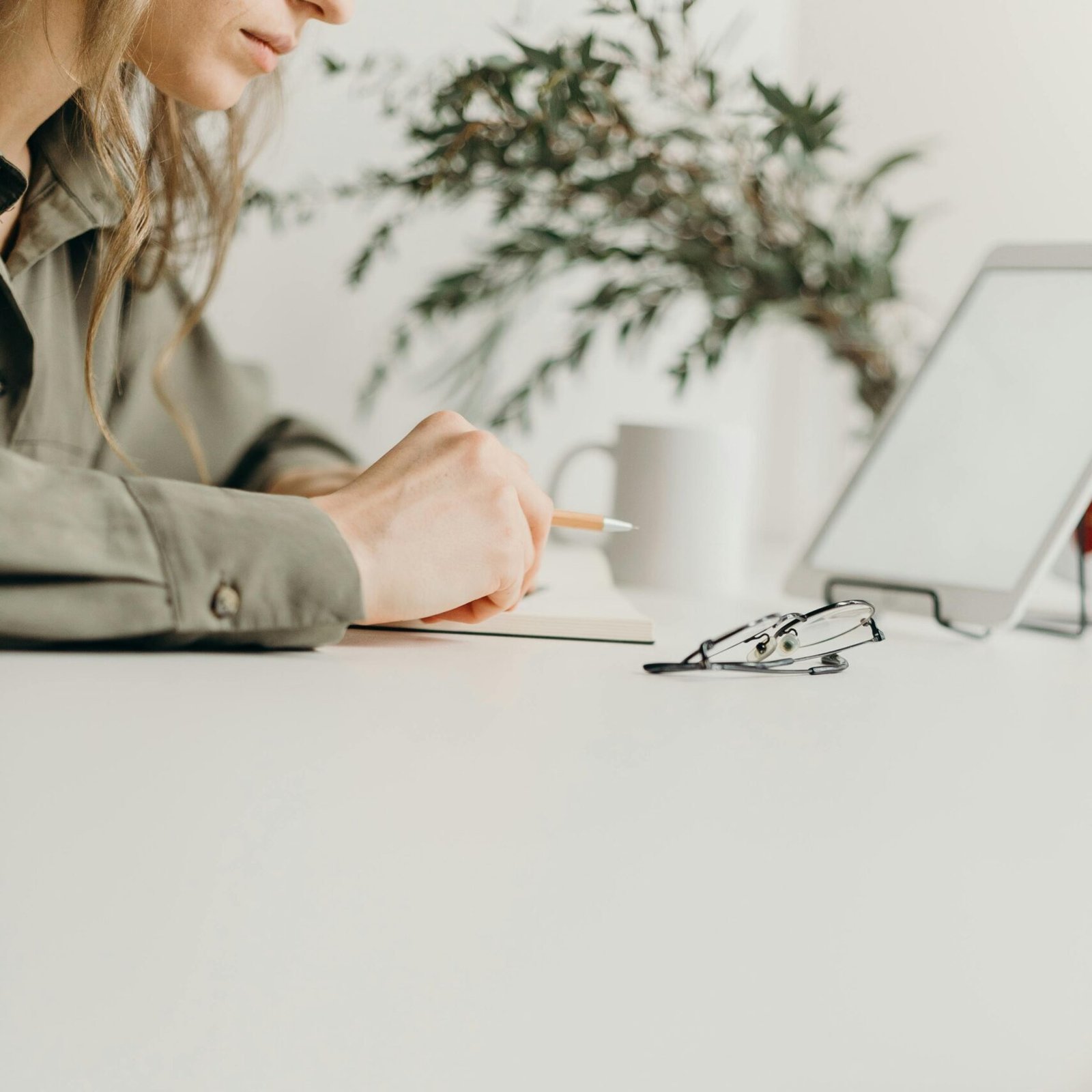 Someone writing in a notebook at a white desk beside a tablet, a cup, and a pair of glasses
